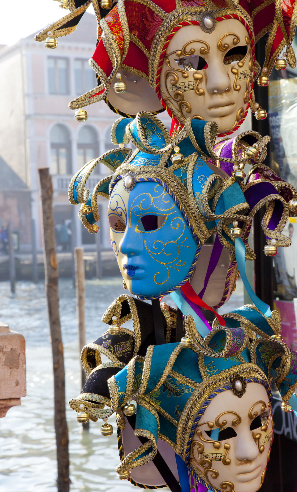 Decorative Venetian masks on display, featuring elaborate designs and intricate details. The background shows a Venetian canal with traditional buildings