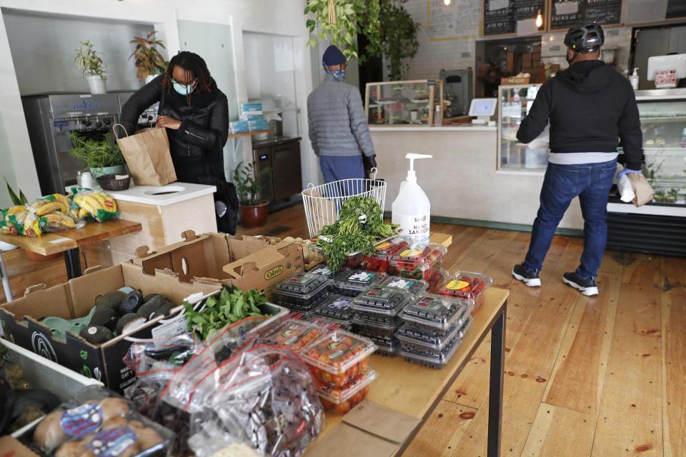 A customer bags her groceries as others wait to pay after shopping for fresh produce, bread and other items, like protective gloves now available at Clementine Bakery in the Brooklyn borough of New York, during the coronavirus outbreak, Thursday, April 16, 2020, in New York. The popular neighborhood spot removed cafe tables and replaced them with a larger table to display fresh fruits and vegetables for sale at a time when customers want those items but don't want to risk standing in line in a crowded grocery store to get them. (AP Photo/Kathy Willens)