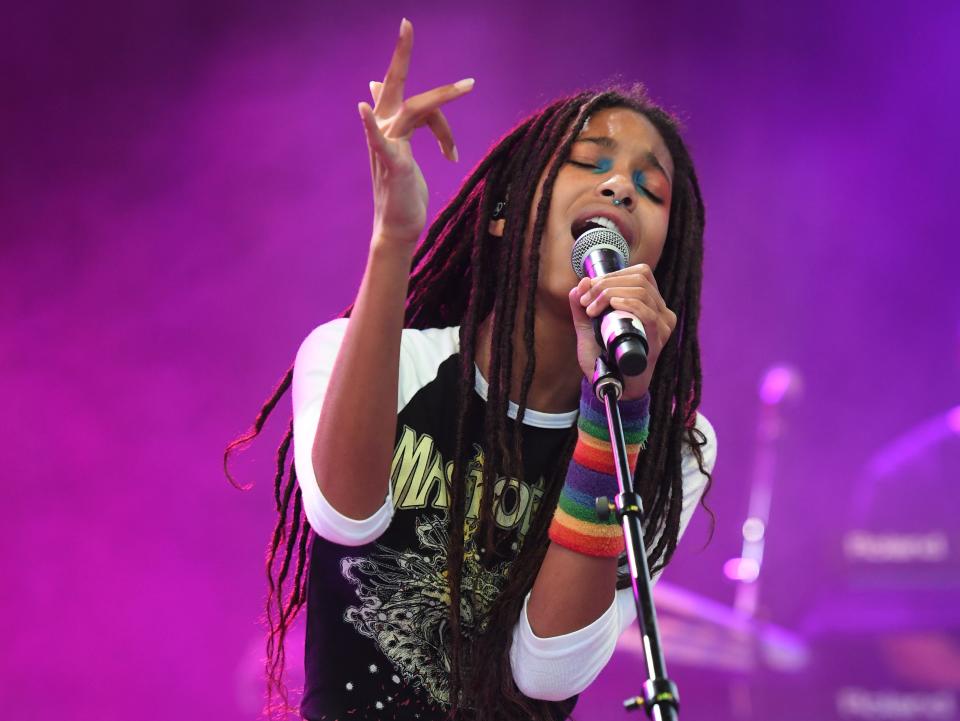 Willow Smith in jeans and a white and black 3/4 sleeve top. She's using a peach and pink scarf as a belt. The lighting behind her is purple while she grips and sings into a mic.