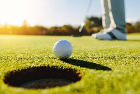 Golf ball approaching hole with putter's shoes and club in the background