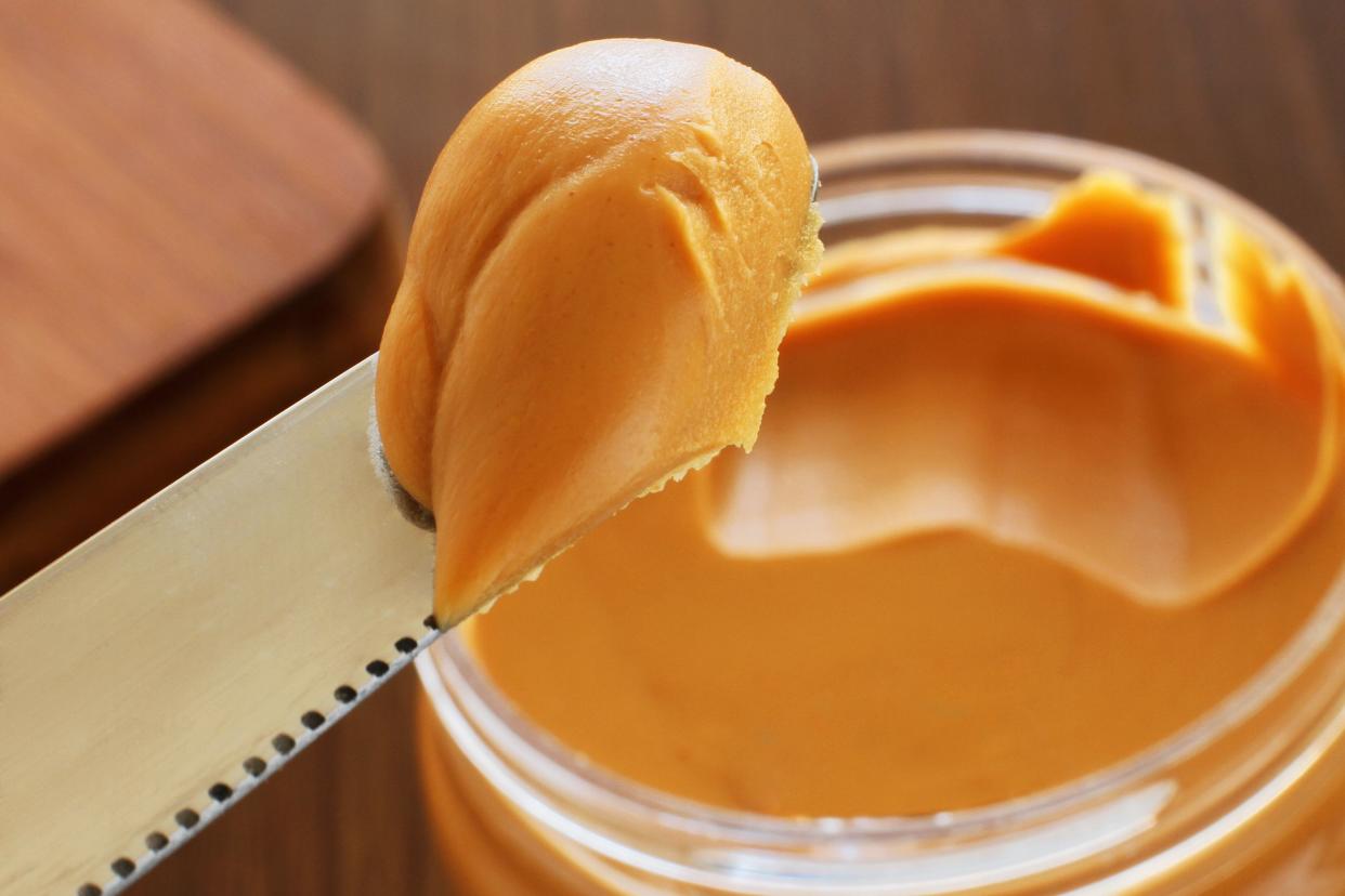 Closeup of a butter knife with a scoop of peanut butter over a blurred full jar of peanut butter with a wooden table and wooden cutting board in the background