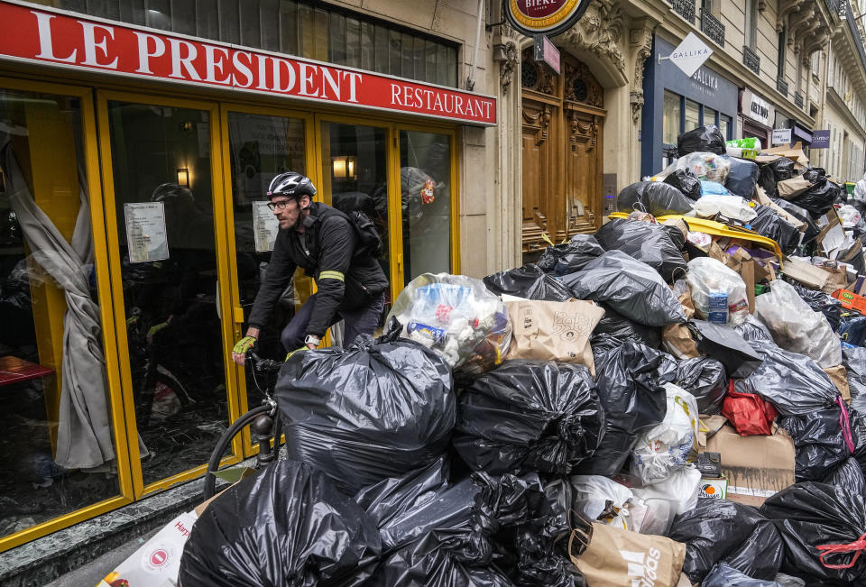 A cyclist rides past an uncollected garbage pile next to the cafe "The President" in Paris, Tuesday, March 21, 2023. The bill pushed through by President Emmanuel Macron without lawmakers' approval still faces a review by the Constitutional Council before it can be signed into law. Meanwhile, oil shipments in the country were disrupted amid strikes at several refineries in western and southern France. (AP Photo/Michel Euler)