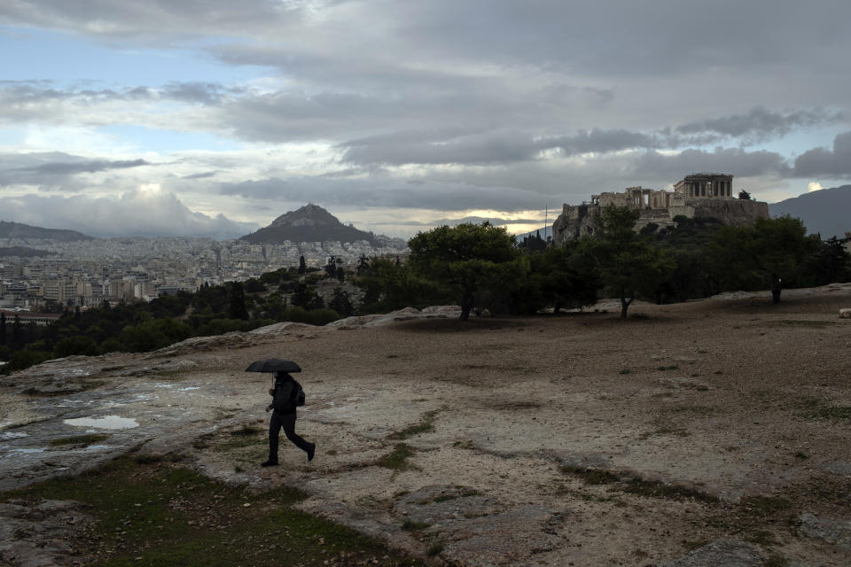 A man with an umbrella walks during a rainfall on Pnyx hill in front the ancient Acropolis hill, with the ruins of the fifth century BC Parthenon temple, in Athens, on Monday, Nov. 30, 2020 Greek officials say the number of new infections is waning in most parts of the country, which has been in lockdown for three weeks.(AP Photo/Petros Giannakouris)