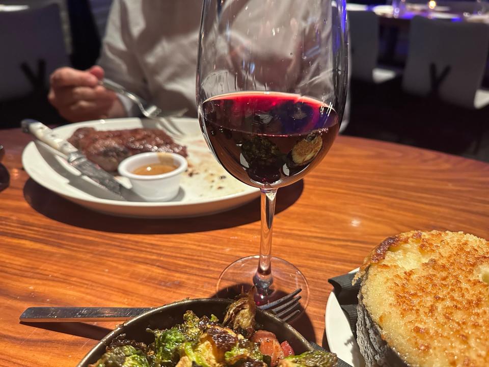 A glass of red wine with a plate of roasted Brussels sprouts, a bowl of mashed potatoes, and a plate of strip steak in the background with someone holding a fork