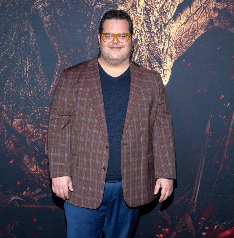 josh gad, a man stands smiling and looking at the camera, he wears a blue tshirt and jeans with a brown jacket and orange glasses