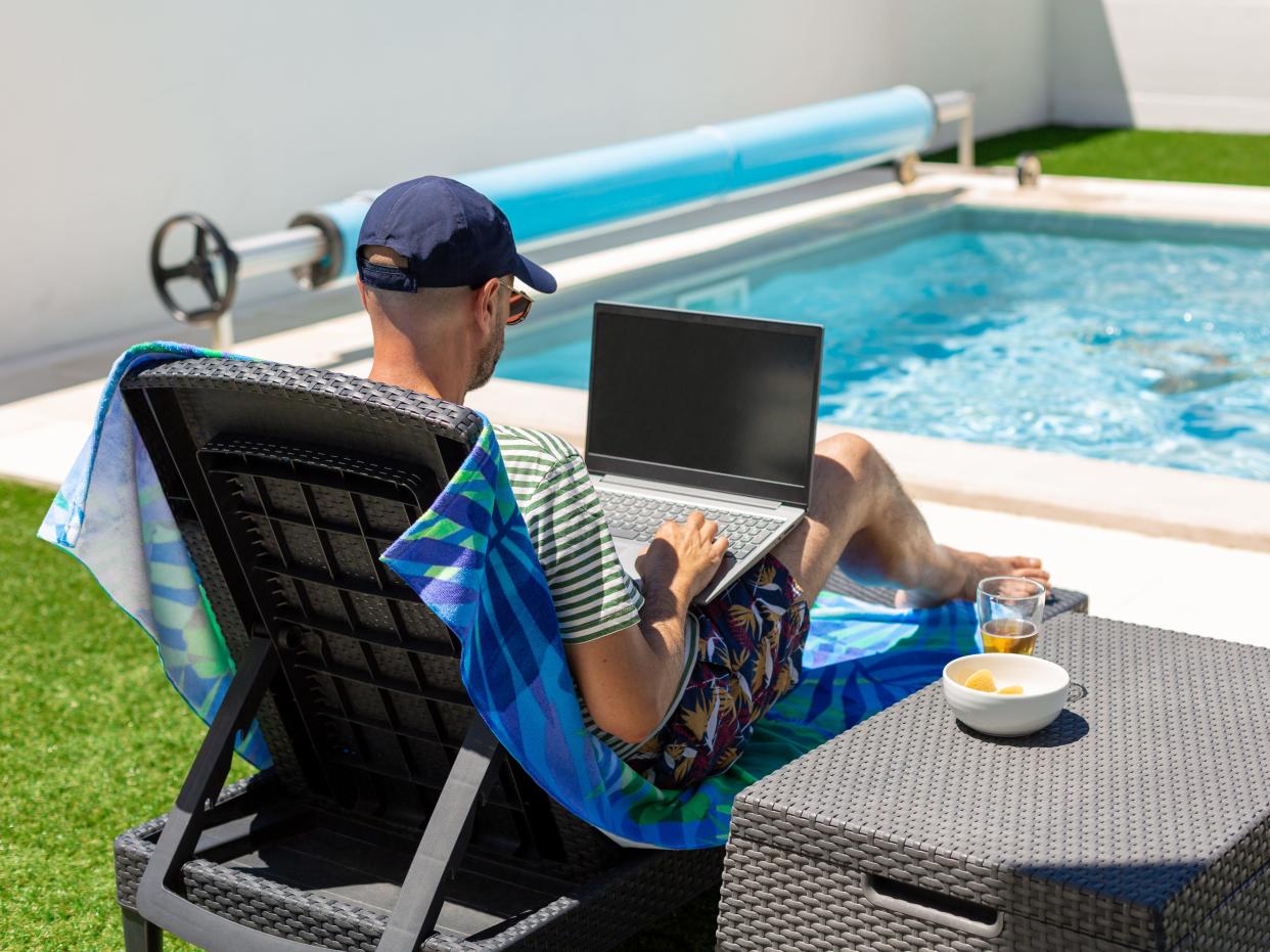 A dad works on his laptop while his daughter swims in a pool.