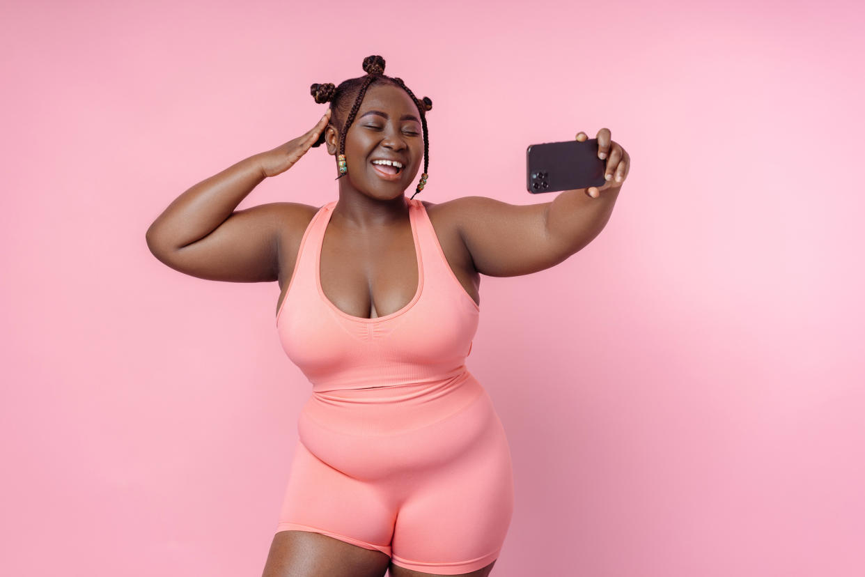 Smiling African woman takes selfie holding phone on pink background. Plus size fashion model poses for pictures, body positive concept.