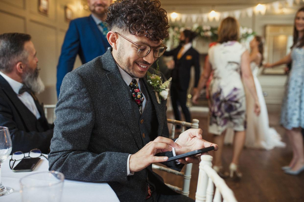 Man at a wedding reception dressed in a suit sitting at a table looking down on his phone smiling while other guests mingle around him.