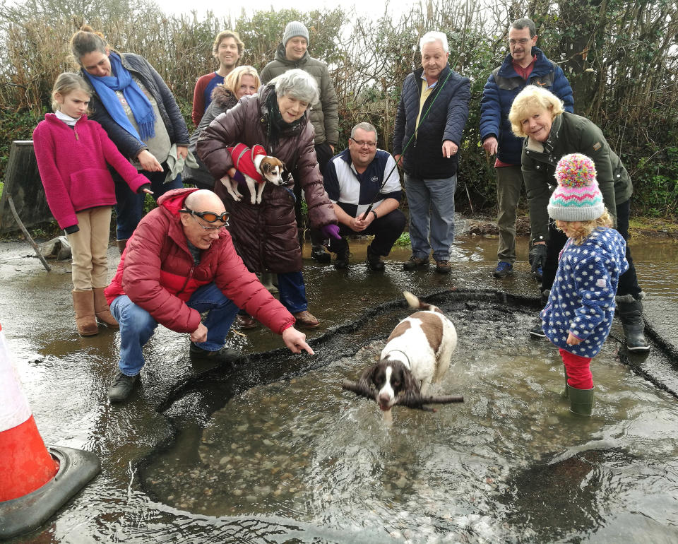 The town councillors and friends in and around the giant pothole in Buckfastleigh, Devon; See SWNS story SWPLpotholes; A group of councillors have dressed up with fishing rods and swimming goggles around an enormous pothole to have a chuckle at the state of Devon's roads. Members of Buckfastleigh Town Council are photographed huddled around the massive hole on the outskirts of the Devon town. There are eight town councillors in the photo, one of whom is dangling the fishing rod inside the pothole while another has donned a pair of swimming goggles. A ninth is behind the camera taking the photo. The water-filled pothole is so big there is a dog with a stick and a young child standing inside.