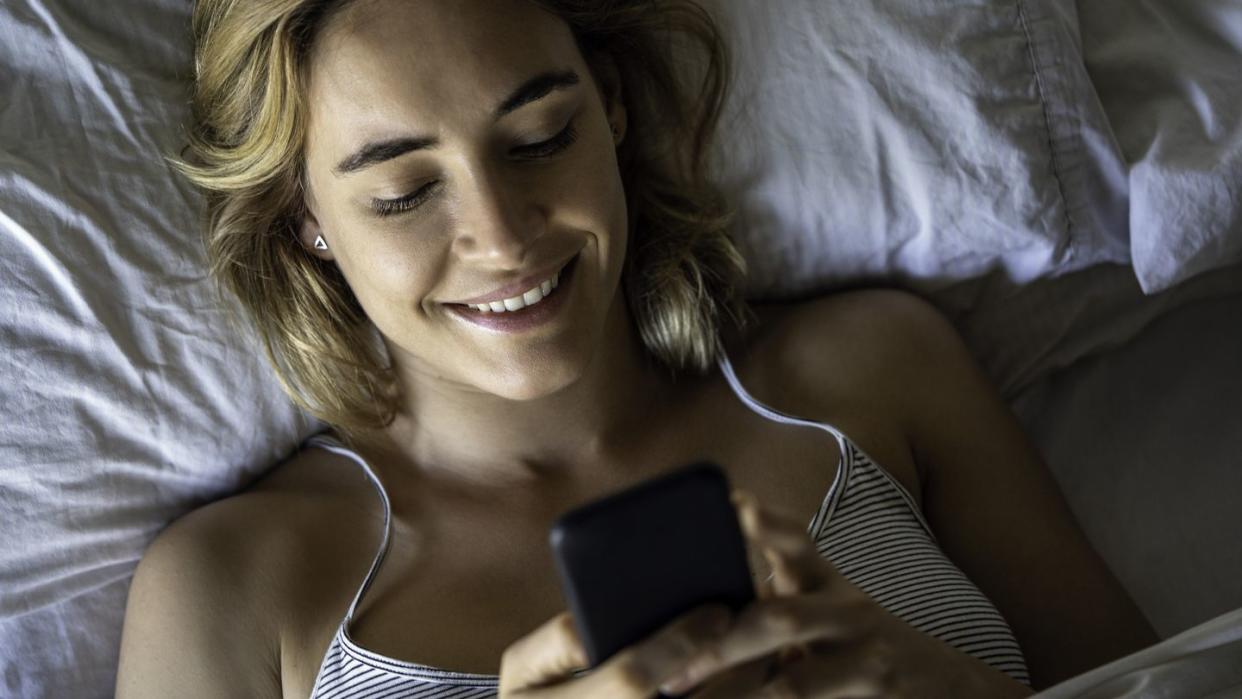 close up of smiling young woman using smartphone while lying on bed in bedroom