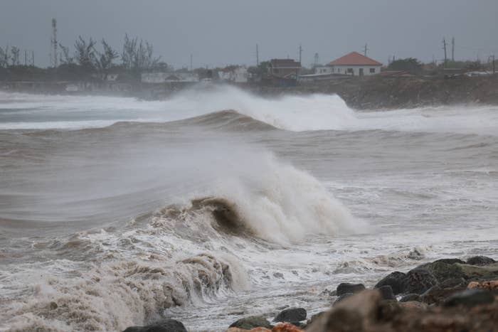 Large waves crash onto a rocky shore with houses in the background, indicating a stormy sea with rough weather conditions