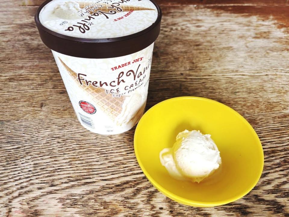 A white carton of vanilla ice cream with an image of a vanilla ice-cream cone and Trader Joe's logo on it. The carton sits next to a yellow bowl holding a scoop of vanilla ice cream