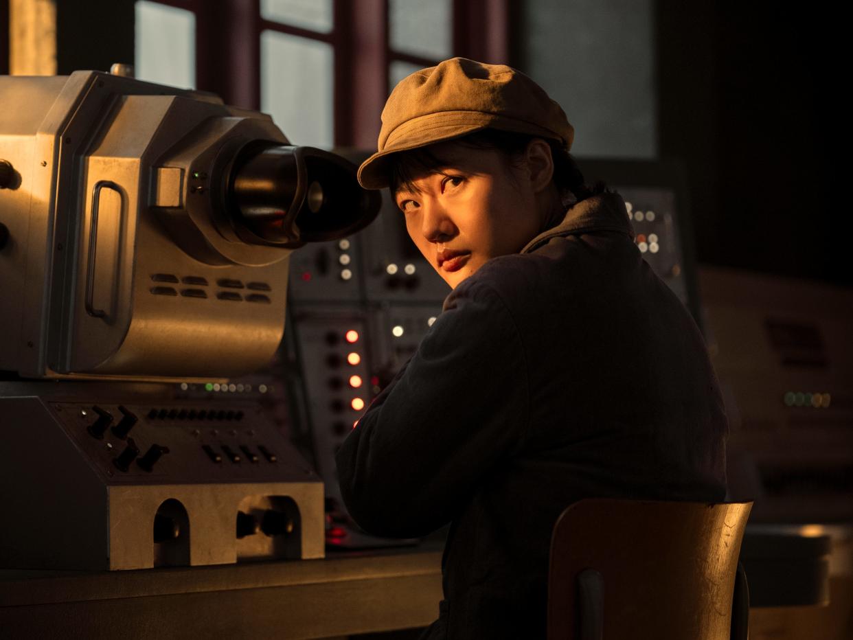 zine tseng as ye wenjie in three body problem. she's a young woman sitting in front of a control panel, wearing a brown hat with a brim, and a blue jacket. bathed in warm light, she looks over her shoulder