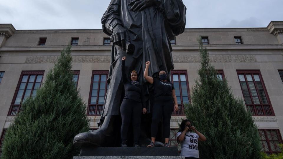 two protestors holding their arm in the air in front of a metal statue of christopher columbus