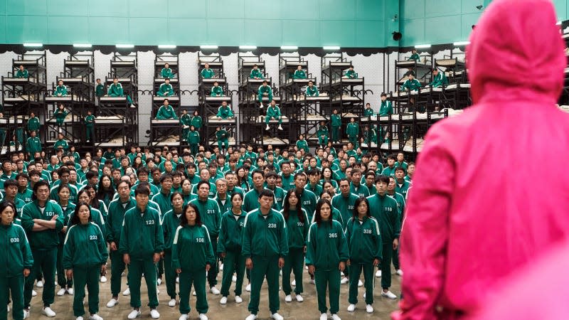 A pink Squid Game guard looks out over a crowd of green, jumpsuit-wearing contestants.