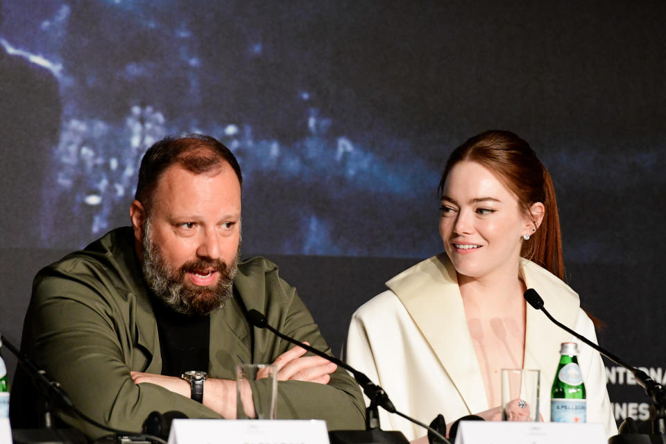 Yorgos Lanthimos and Emma Stone sit at a press conference panel. Yorgos speaks into a microphone while Emma smiles looking at him