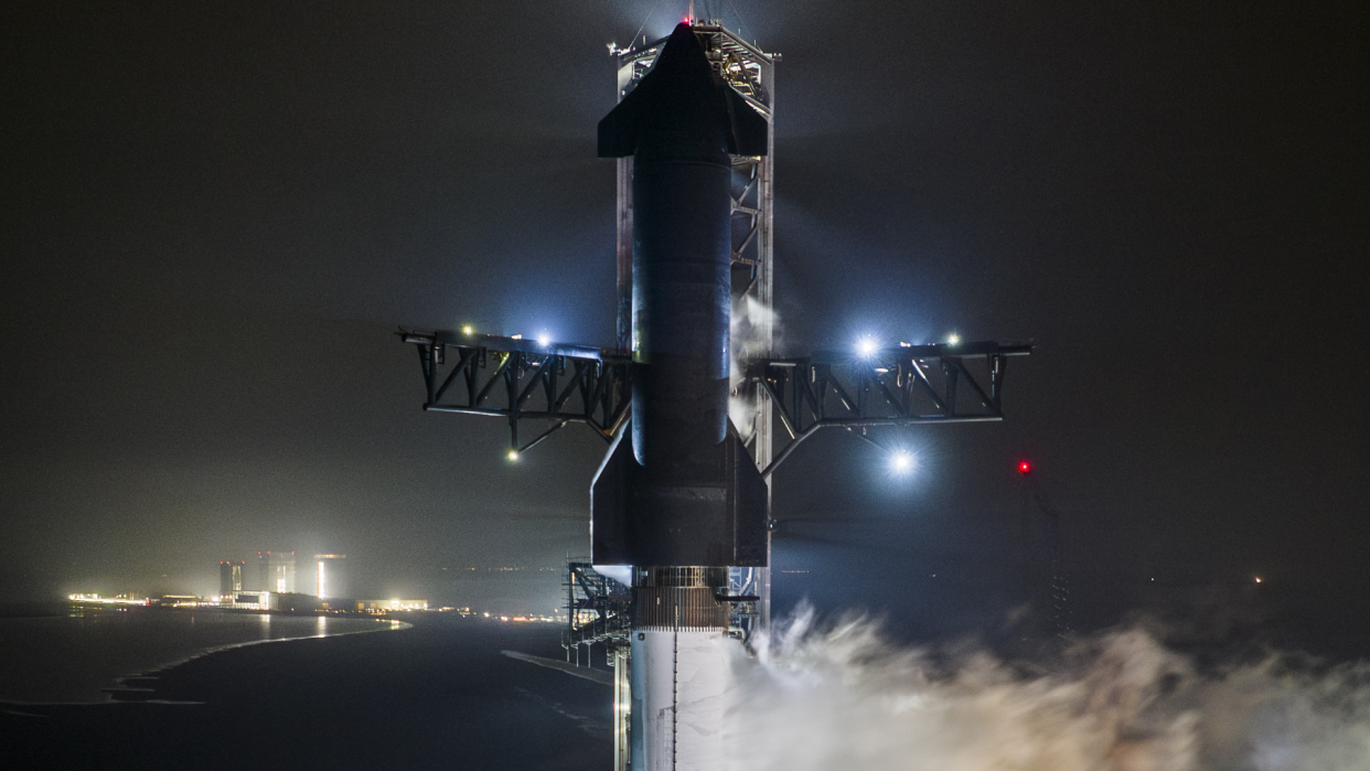  A large black and silver rocket vents vapor at night while standing upright on a launch pad. 