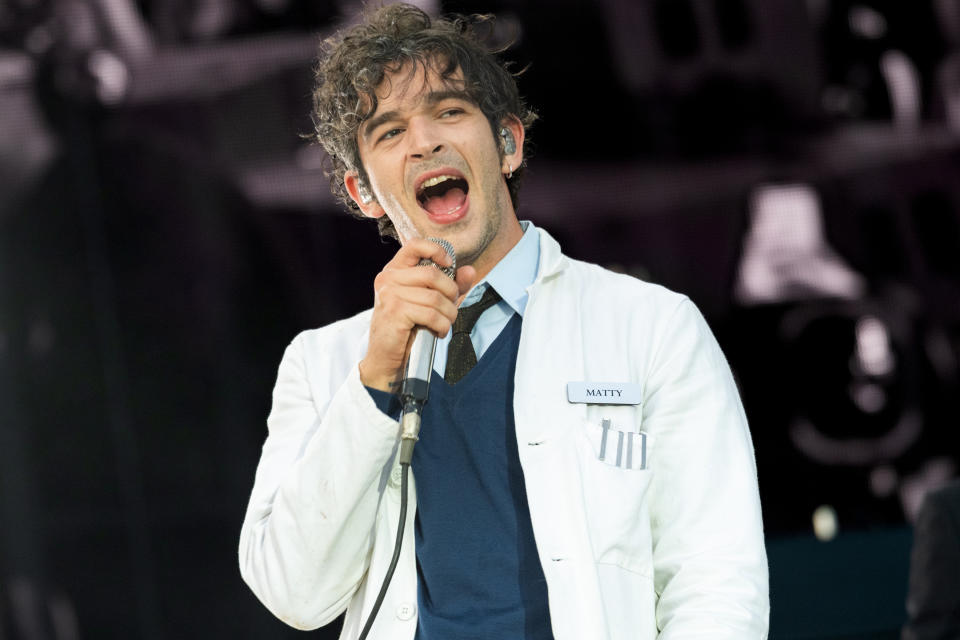 Matty Healy of The 1975 performing on stage, wearing a white lab coat with "Matty" name tag