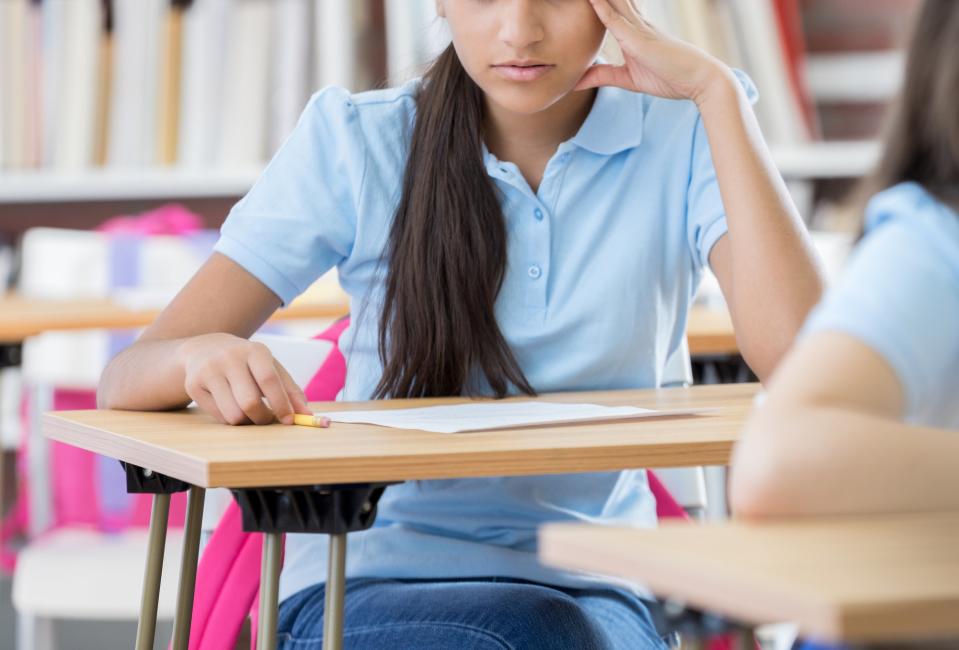 Two students, one focused on a test and the other looking down, sit at desks in a classroom with bookshelves in the background