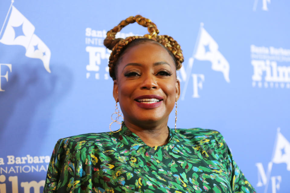 Aunjanue smiling at event, styled in a patterned dress with hoop earrings, hair in a braided bun