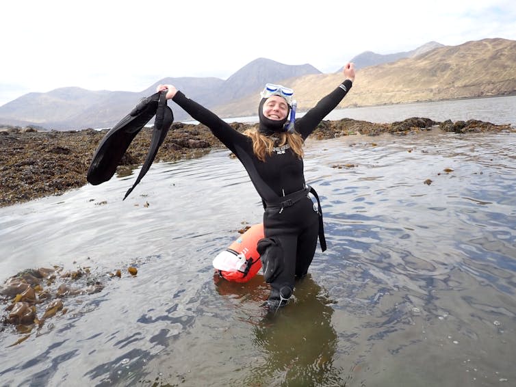 Woman in black wetsuit and snorkel mask, standing in shallow water with orange tow float, arms wide open and holding finds in her hands, rocky coast behind her