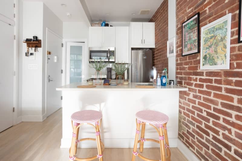 Two red and white bistro bar stools in front of a white bar countertop in an all white kitchen with brick walls and stainless steel appliances.