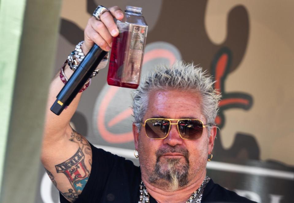 Guy Fieri wears sunglasses and toasts with a drink and a microphone in his hand.