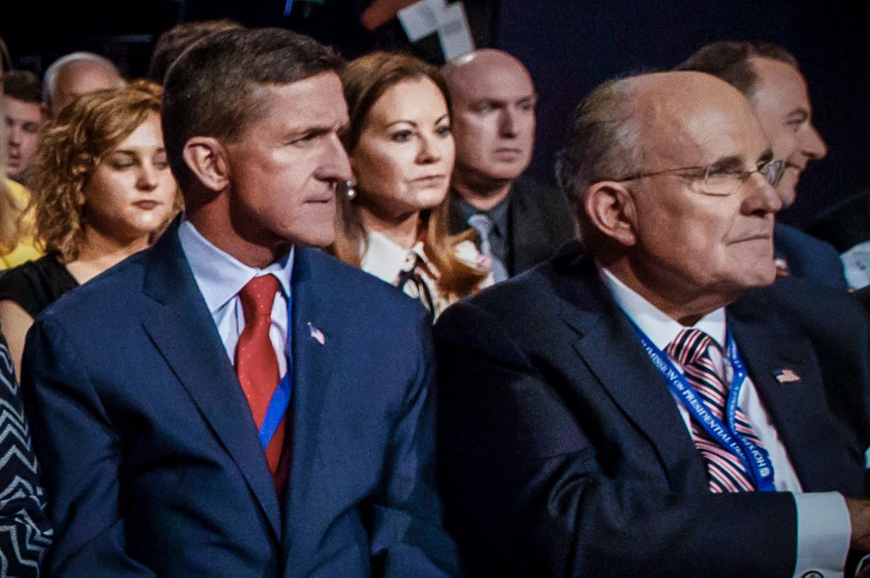 Left: Michael Flynn seated in a crowd in a blue suit and a red and a blue tie; Right: Rudy Giuliani seated next to him in a striped red and black tie and a black suit