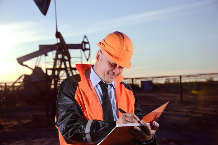 A man writing in a notebook with an oil well behind him