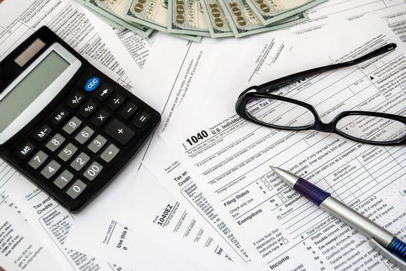 Tax forms, cash, a calculator, glasses, and a pen spread out on a surface.