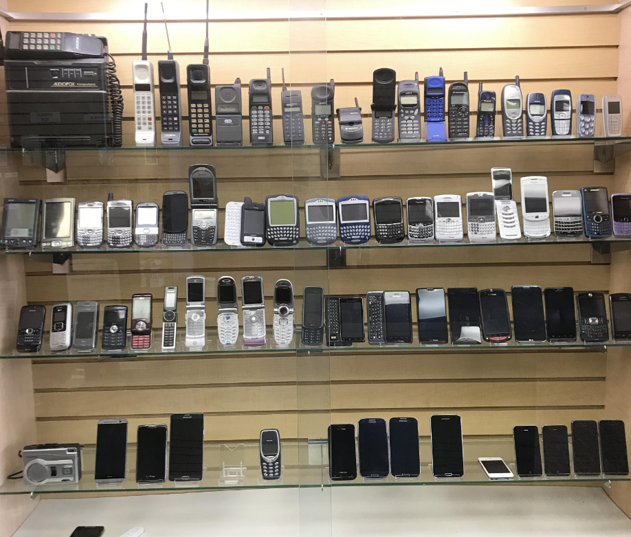 A display case with five shelves of various models of old mobile phones, organized by model and design, ranging from early brick phones to early smartphones