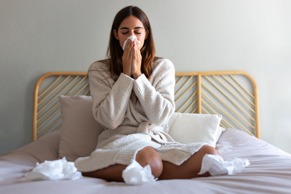 Young caucasian woman blowing nose with paper tissue wearing a robe sitting on bed suffering from a cold. Cold and sickness concept. what is a syndemic