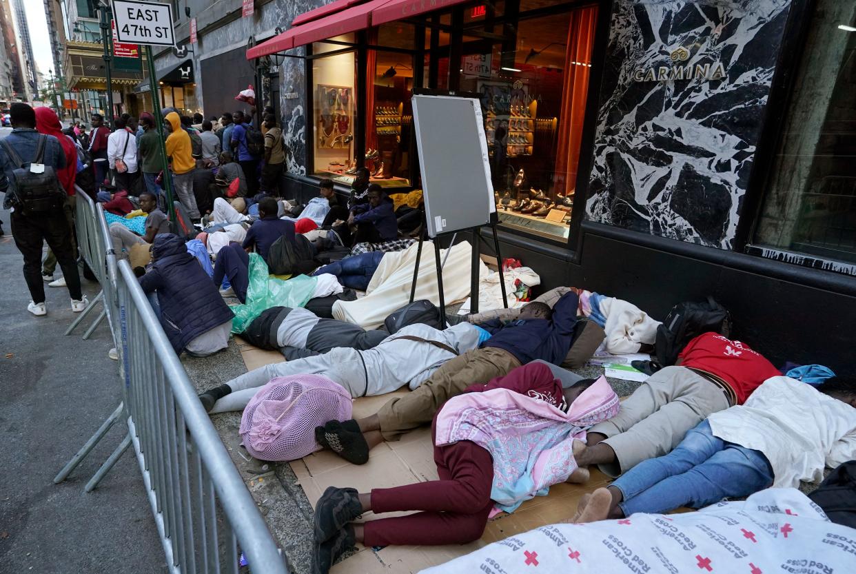 A row of migrants, protected behind a metal barricade, sleep on cardboard on the sidewalk.