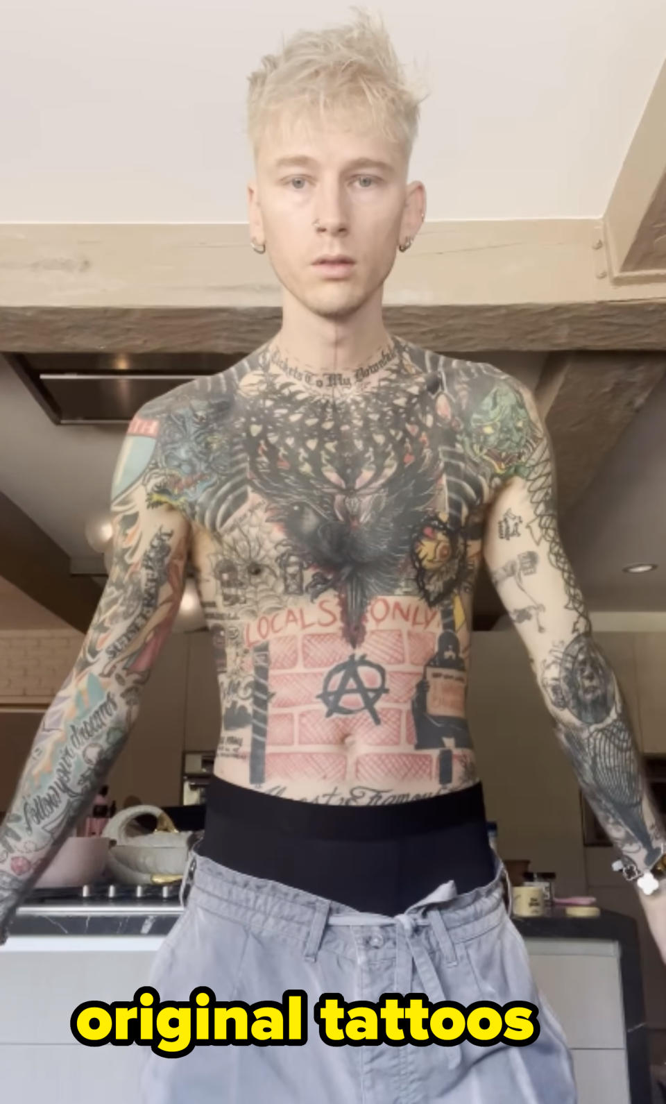 Man with tattoos on torso and arms, standing in a kitchen