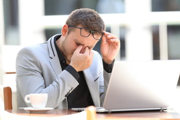 A businessman, sitting at a bistro table with his laptop and some coffee, lifts his glasses to rub his eyes.