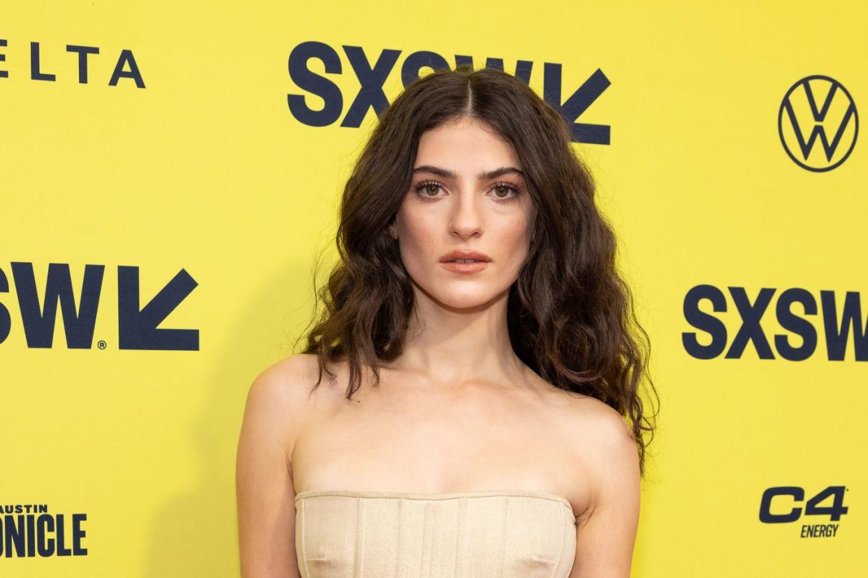 ella rubin, a young woman stands looking at the camera with a neutral facial expression, she has long brown hair worn in loose waves and wears a strapless beige dress