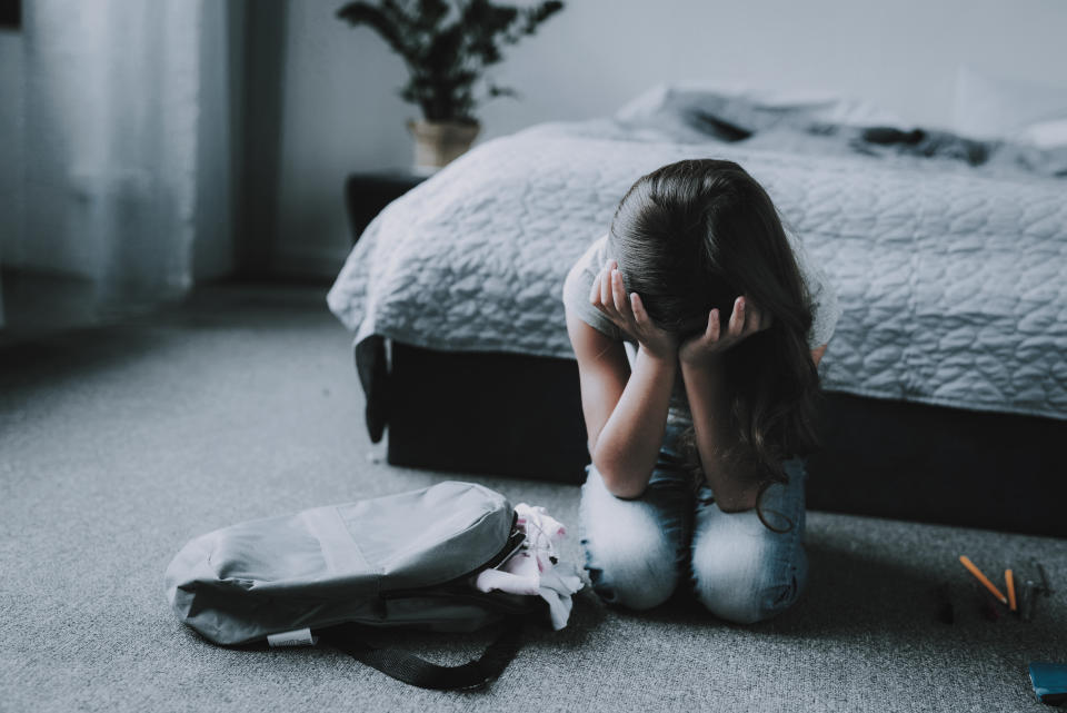 Girl Sitting on Floor in Bedroom and Crying. Portrait of Unhappy Black-Haired Child with Closed Eyes Covers Face with Hands Wearing Gray T-Shirt Sits Near Backpack next to Bed in Modern Apartment