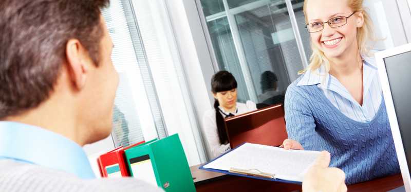 Smiling woman hands paperwork to a man over a desk.