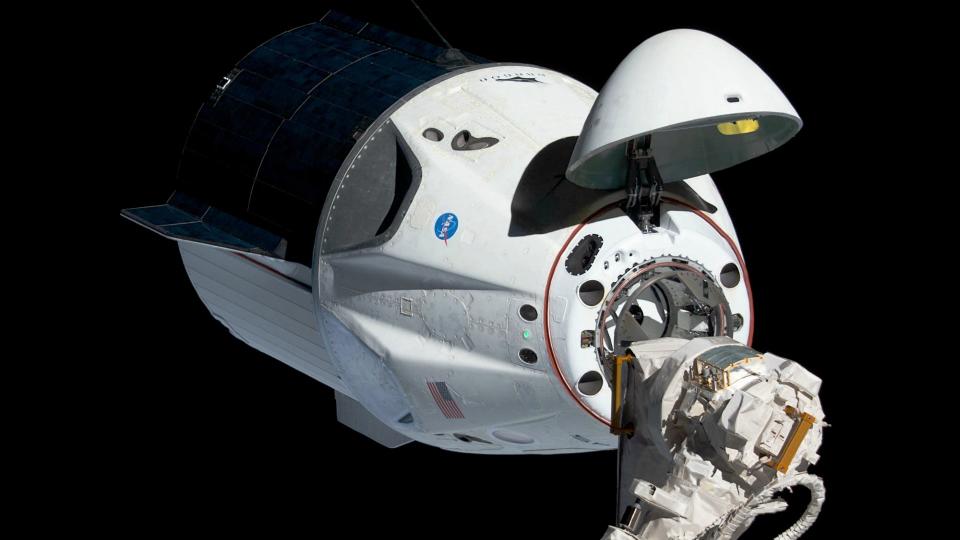 a cone-shaped spacecraft with hatch open in space. it is next to the top of a robotic arm