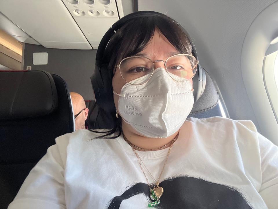 Author Libby Torres sits in a window seat on an American Airlines flight wearing a white KN95 face mask, gold wire-rimmed glasses, black headphones, a white T-shirt, and gold jewelry.