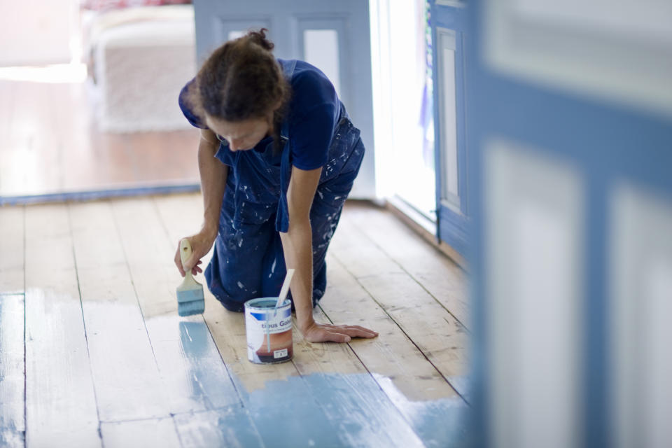 Person kneeling while applying paint to a wooden floor using a roller brush; paint can beside them