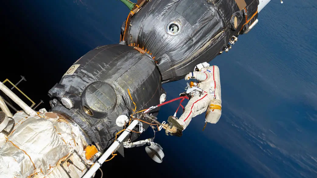  A cosmonaut in an eva suit floats outside the space station near a soyuz vehicle, feet secured to an extended mechanical arm. Russian spacewalker Oleg Kononenko (suit with red stripes) works outside the International Space Station on Dec. 11, 2018. He was inspecting and gathering samples from the Soyuz MS-09 spacecraft following a small pressure leak; the leak had already been sealed from the inside shortly after it was detected. 