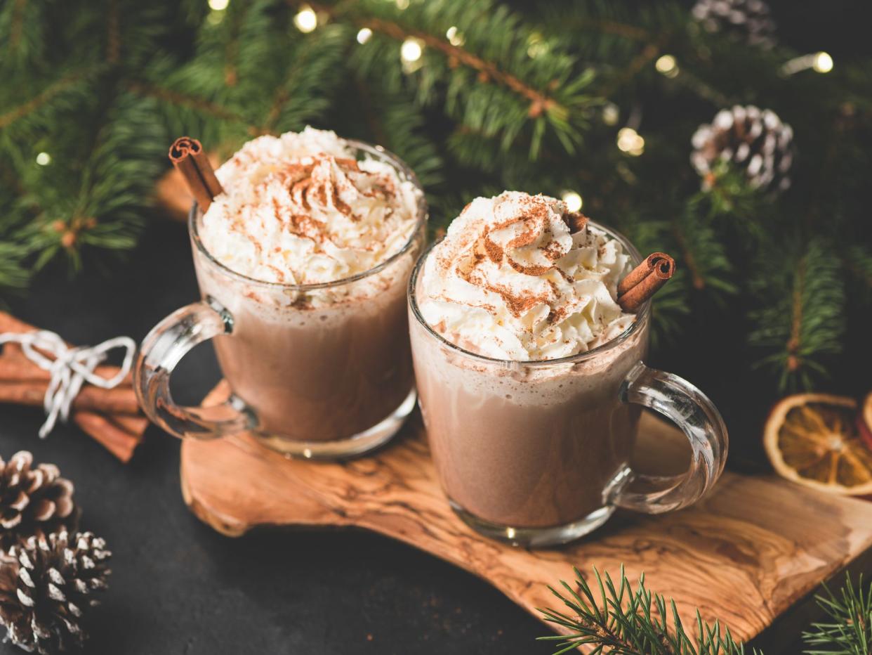 Two cups of hot chocolate with whipped cream and cinnamon on wooden serving board surrounded with fir tree, Christmas lights, cinnamon and pine cones. Cozy Christmas Drink