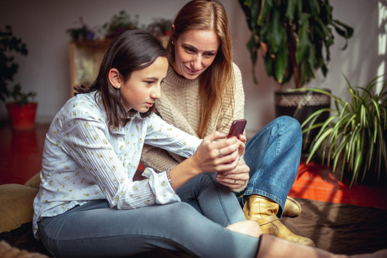 A young girl uses a mobile phone with her mother smiling and watching over her