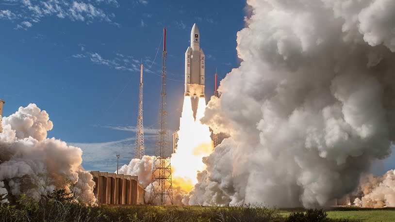 A white Ariane 5 rocket llfts off against a blue sky, with a white cloud of smoke blowing out from the ground below.