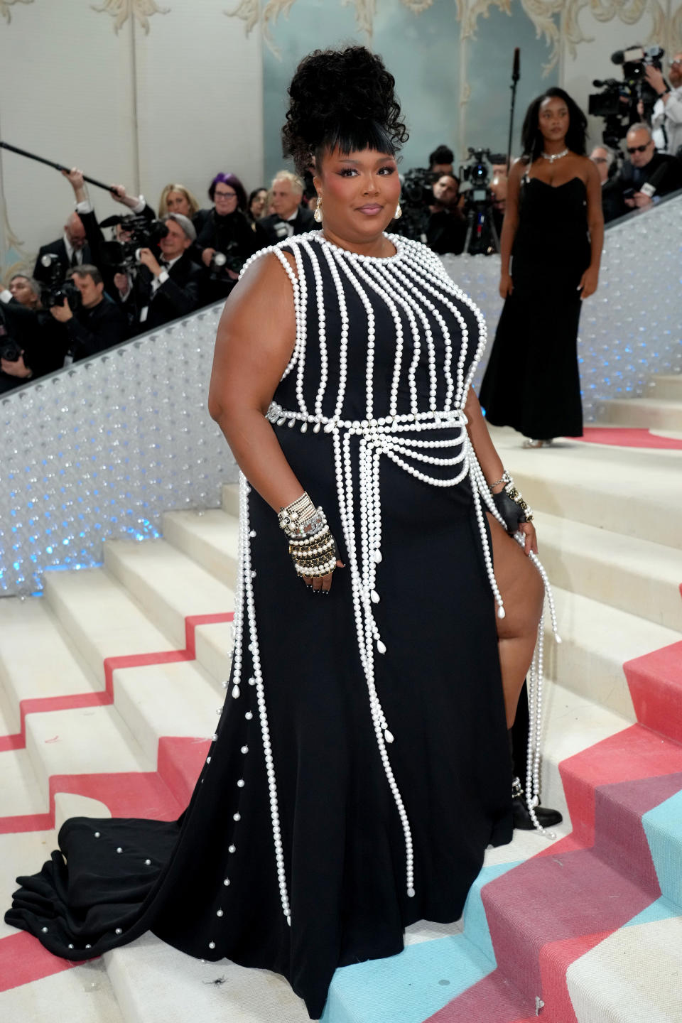 Lizzo stands on stairs in a black dress with white beaded embellishments, bracelets, and updo hairstyle