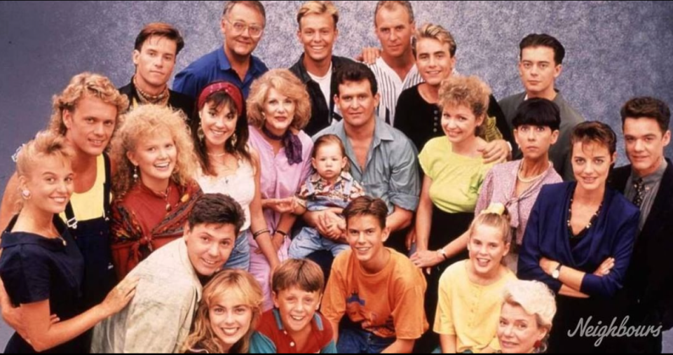 The cast of Aussie soap Neighbours in the late 80s including 22 adults and young people and a baby. They are grouped closely together and smiling at the camera.. 