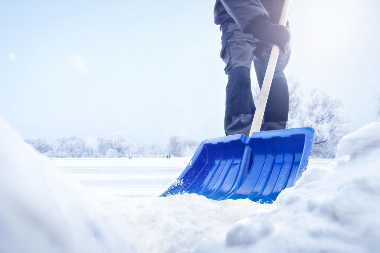 A person using a snow shovel in a snowy landscape. Only the lower part of the person is visible. Low angle shot with snow piles in the foreground.