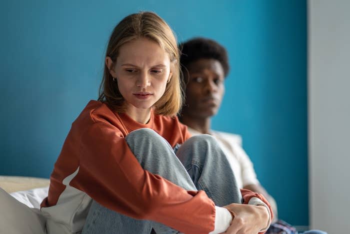 A woman in casual clothing sits on a bed, looking away with a concerned expression. A man is seated in the background, possibly engaged in a serious conversation