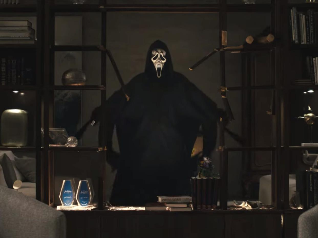 ghost stands in between two bookcases, arms outstretched, in a dimly lit apartment during a scene from the "Scream 6" trailer.